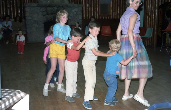 Children in a conga line led by an adult across the wooden floor of the recreation hall, stone fireplace behind them